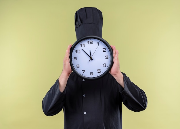 Cuoco unico maschio che indossa l'uniforme nera e cappello da cuoco che tiene l'orologio da parete che nasconde il suo volto dietro in piedi su sfondo verde