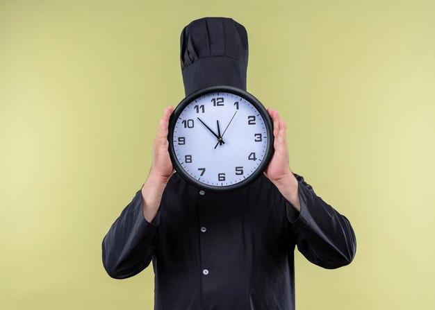 Male chef cook wearing black uniform and cook hat holding wall clock hiding his face behind it standing over green background