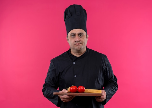 Male chef cook wearing black uniform and cook hat holding wooden cutting board with tomatoes looking at camera with serious face standing over pink background