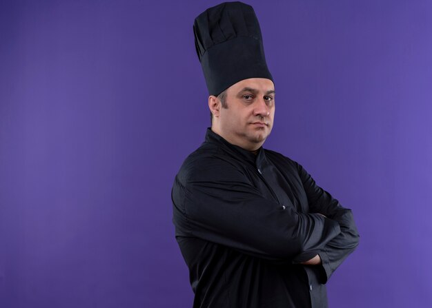 Male chef cook wearing black uniform and cook hat looking at camera with confident expression with crossed arms on chest standing over purple background