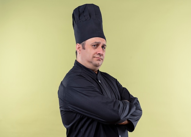 Foto gratuita cuoco unico maschio che indossa l'uniforme nera e cappello da cuoco che guarda l'obbiettivo con espressione seria fiduciosa con le braccia incrociate sul petto in piedi su sfondo verde