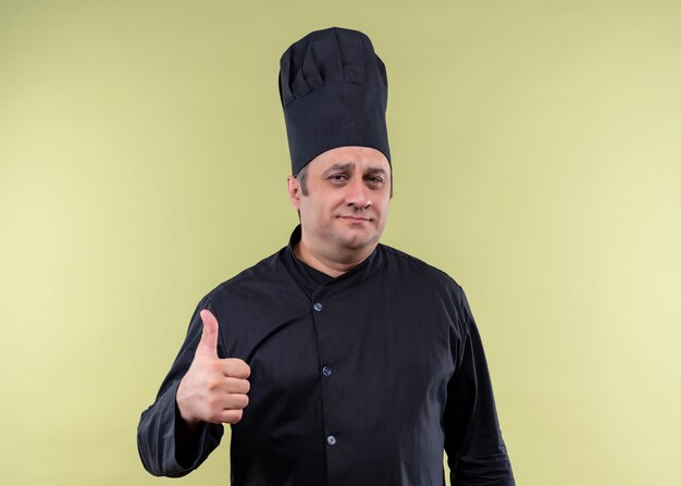 남성 요리사는 검은 제복을 입고 요리하고 녹색 배경 위에 서있는 엄지 손가락을 보여주는 얼굴에 미소로 카메라를보고 모자를 요리합니다.