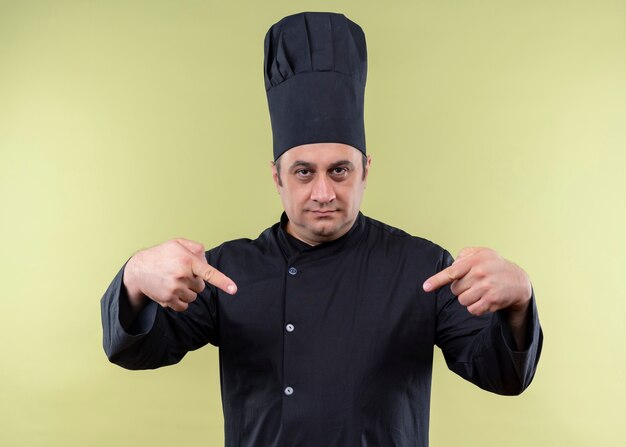 남성 요리사는 검은 색 유니폼을 입고 요리사 모자를 요리하고 녹색 배경 위에 서있는 자신을 손가락으로 가리키는 자신감을 찾고 있습니다.