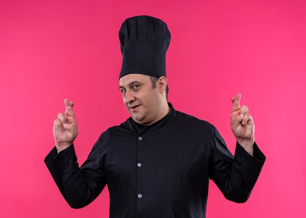 남성 요리사는 검은 색 유니폼을 입고 요리사 모자를 만들고 분홍색 배경 위에 서있는 손가락을 건너는 바람직한 소원을 만듭니다.