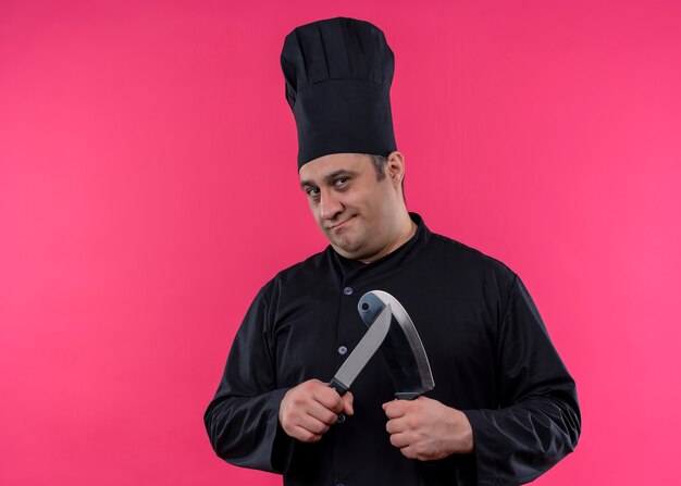 Шеф-повар-мужчина в черной униформе и поварской шляпе точит ножи, глядя в камеру с серьезным лицом, стоящим на розовом фоне