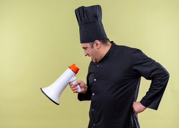 Male chef cook wearing black uniform and cook hat shouting to megaphone standing over green background