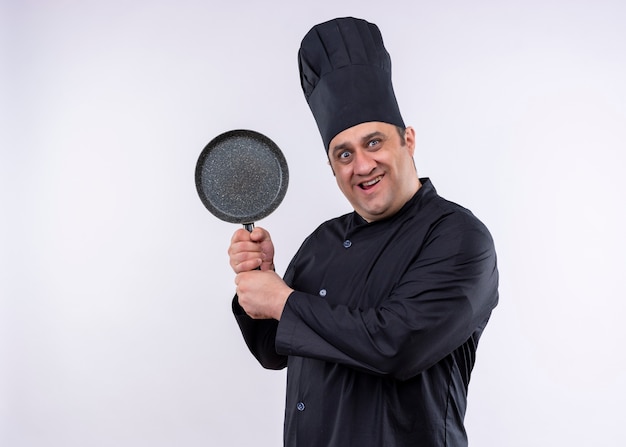 Cuoco unico maschio che indossa l'uniforme nera e cappello da cuoco che mostra pan loking alla fotocamera in piedi sorpreso ed eccitato su sfondo bianco