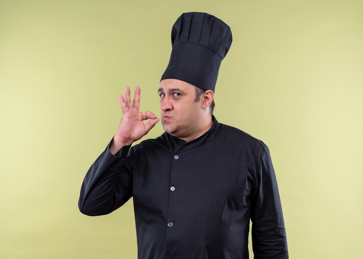Free Photo male chef cook wearing black uniform and cook hat showing sign for delicious standing over green background