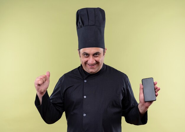 남성 요리사는 검은 색 유니폼을 입고 요리하고 녹색 배경 위에 서있는 주먹을 떨리는 스마트 폰을 보여주는 모자를 요리합니다.