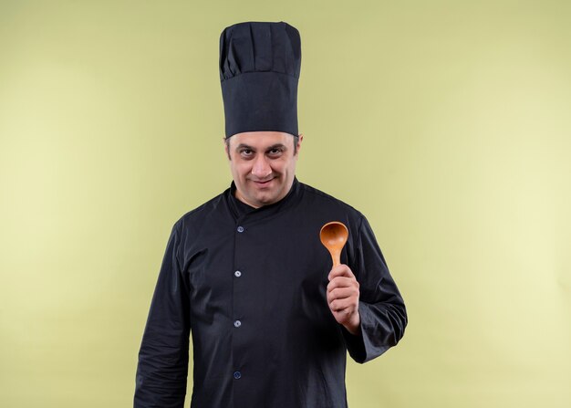 Male chef cook wearing black uniform and cook hat showing wooden spoon looking at camera with smile on face standing over green background