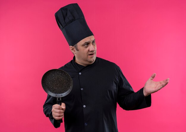 Male chef cook wearing black uniform and cook hat standing over pink background