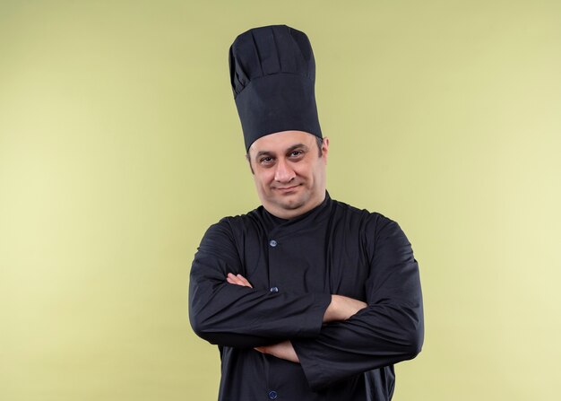 Male chef cook wearing black uniform and cook hatlooking at camera with crossed hands looking confident standing over green background