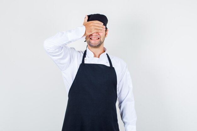 Male chef covering eyes with hand in uniform, apron and looking excited. front view.