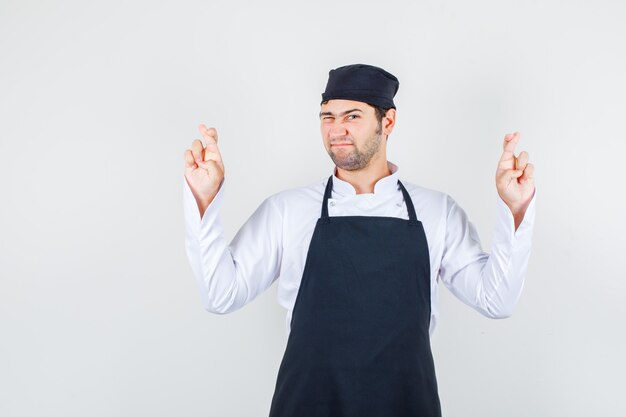 Male chef crossing fingers with blinked eye in uniform, apron , front view.