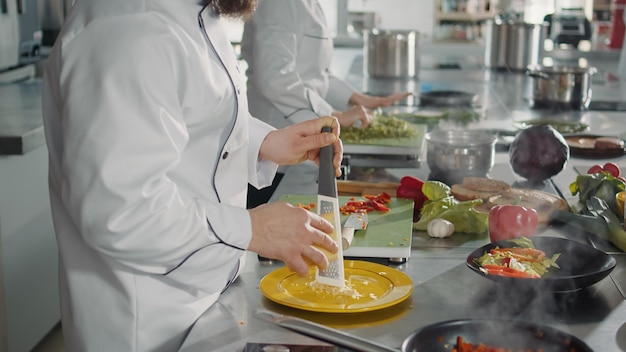 Male chef grating parmesan cheese on kitchen grater to cook gastronomy recipe in professional cuisine. Authentic cook preparing shredded cheddar ingredient to make gourmet meal dish.