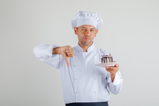Male chef holding cake and showing thumb down in uniform, apron and hat front view.