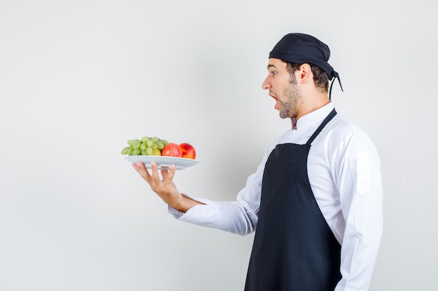 Male chef holding fruits in plate in uniform, apron and looking amazed. front view.