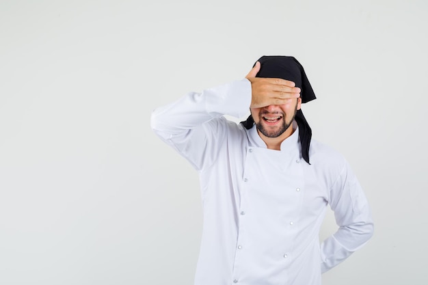 Male chef holding hand on eyes in white uniform and looking eager , front view.