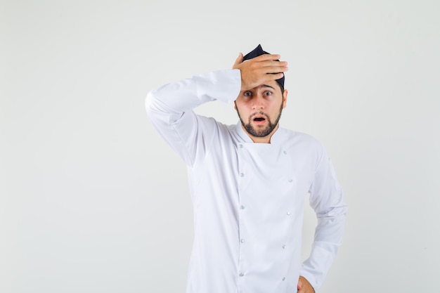 Male chef holding hand on his forehead in white uniform and looking surprised , front view.