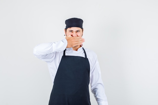 Cuoco unico maschio che tiene la mano sulla bocca in uniforme, grembiule e sguardo irritato. vista frontale.