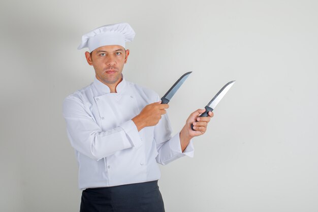 Male chef holding kitchen knives in uniform, hat and apron