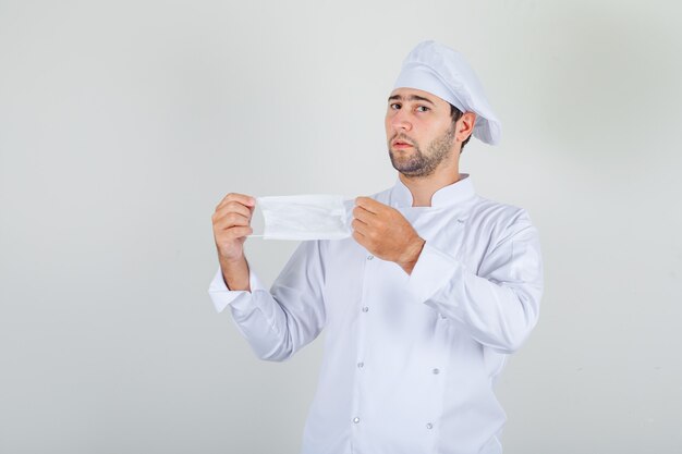 Male chef holding medical mask in white uniform and looking hesitant.