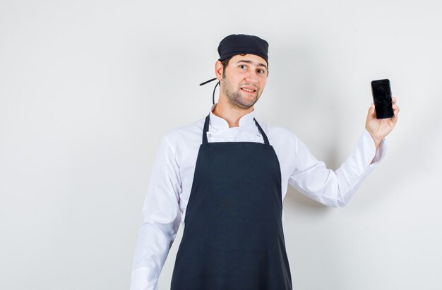 Мужской шеф-повар держит мобильный телефон в униформе, фартуке и выглядит оптимистично. передний план.