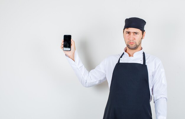 Male chef holding mobile phone in uniform, apron and looking upset , front view.