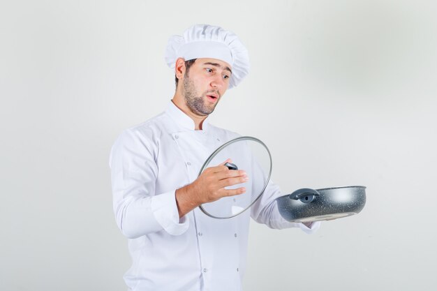 Male chef holding pan and glass lid in white uniform and looking surprised.