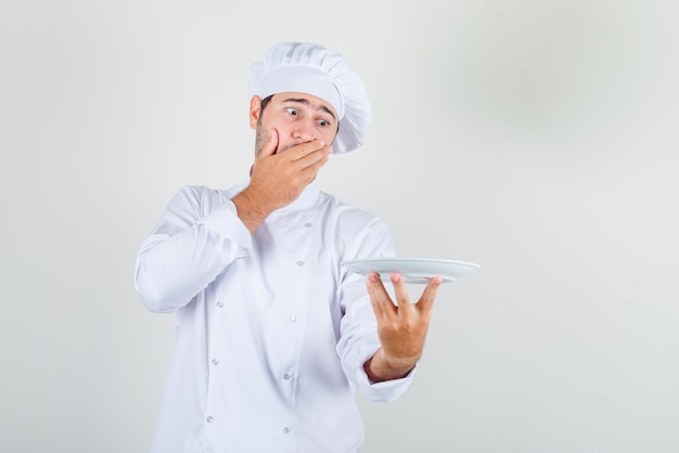 Male chef holding plate with hand on head in white uniform and looking surprised