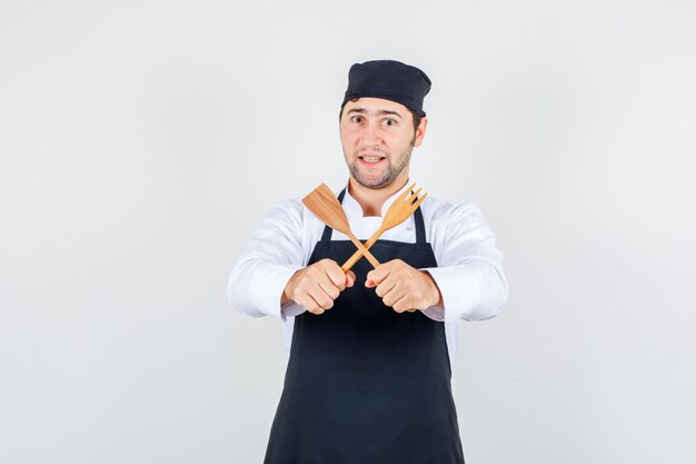 Male chef holding wooden fork and spatula in uniform, apron and looking cheery , front view.