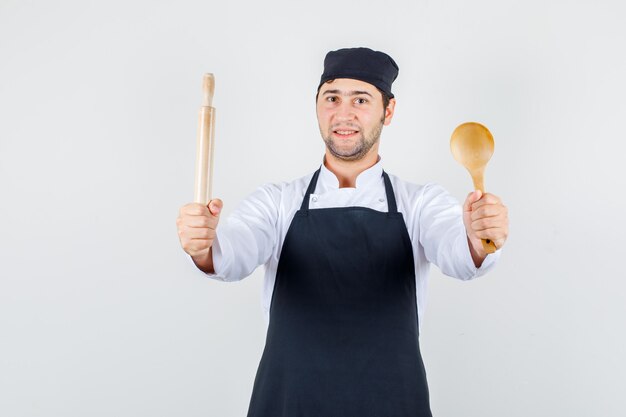 Male chef holding wooden spoon and rolling pin in uniform, apron front view.