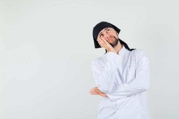 Male chef leaning cheek on raised palm in white uniform and looking thoughtful. front view.