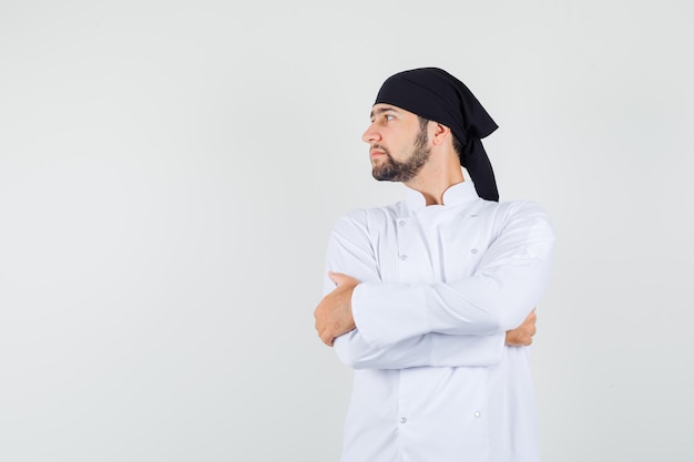 Мужчина-повар смотрит в сторону со скрещенными руками в белой форме и смотрит сосредоточенно, вид спереди.