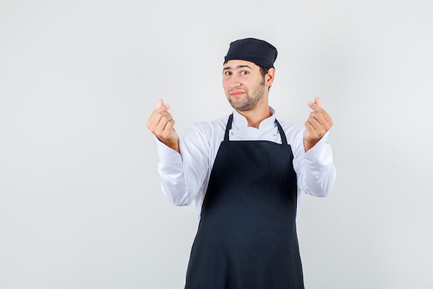 Cuoco unico maschio che fa gesto di amore coreano in uniforme, grembiule e guardando ottimista, vista frontale.