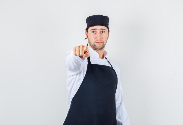 Male chef pointing finger at you in uniform, apron and looking serious , front view.