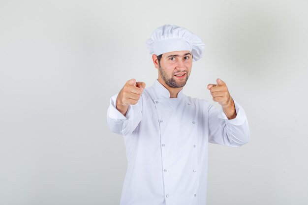 Male chef pointing fingers to camera in white uniform and looking cheerful.