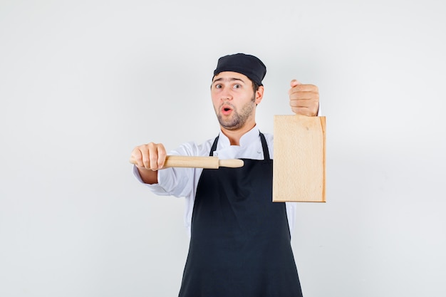 Cuoco unico maschio che indica il mattarello al tagliere in uniforme, grembiule, vista frontale.