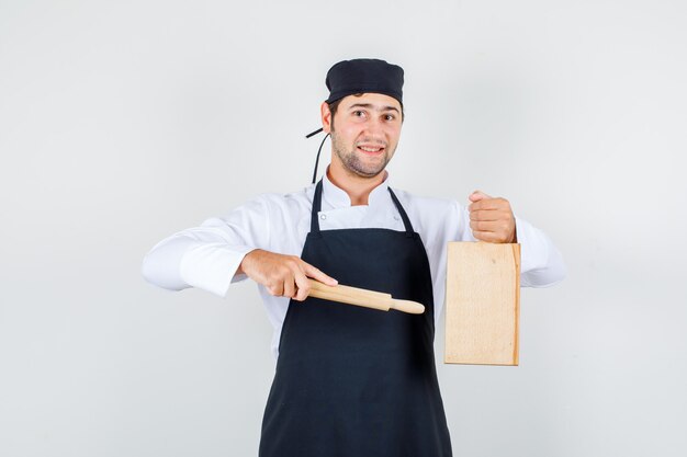 Шеф-повар-мужчина указывая скалкой на разделочную доску в униформе, фартуке и веселый вид, вид спереди.