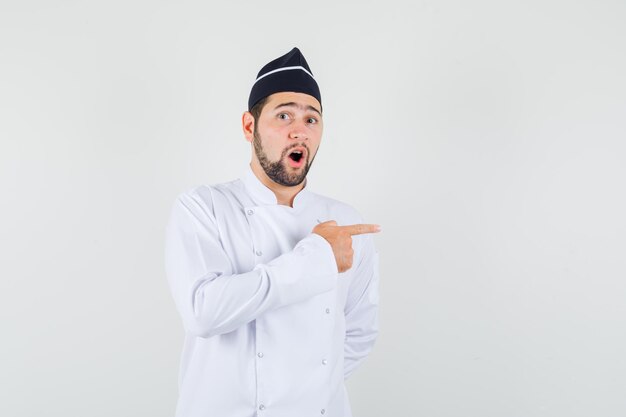 Male chef pointing to the side in white uniform and looking curious , front view.