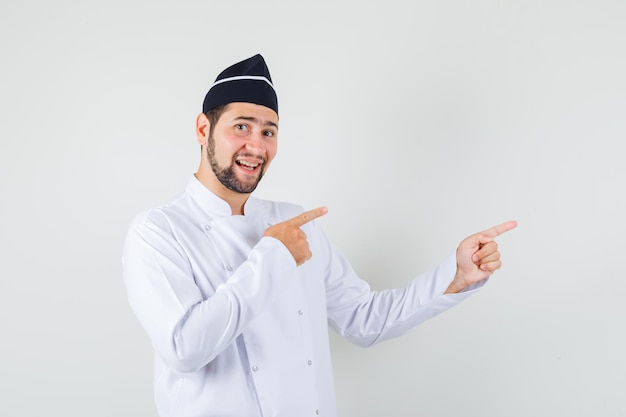 Male chef pointing to the side in white uniform and looking merry , front view.