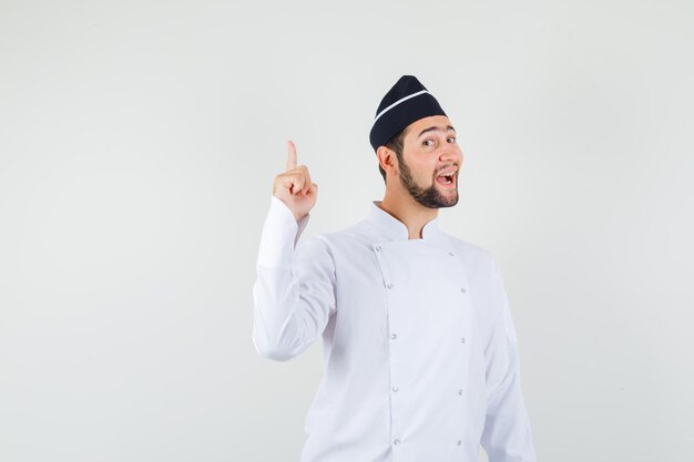 Male chef pointing up in white uniform and looking merry. front view.