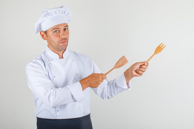 Male chef pointing wooden kitchen utensils away in uniform, apron and hat
