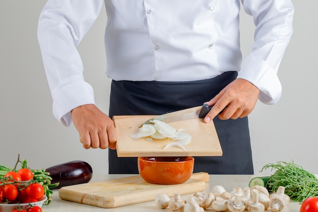 Male chef pouring chopped onions into bowl in uniform and apron in kitchen