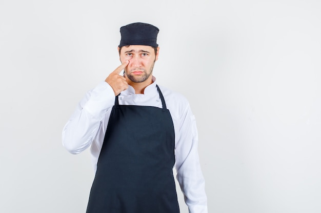 Male chef pulling down eyelid in uniform, apron and looking sad , front view.