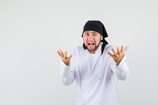 Male chef raising hands in questioning manner in white uniform and looking irritated , front view.