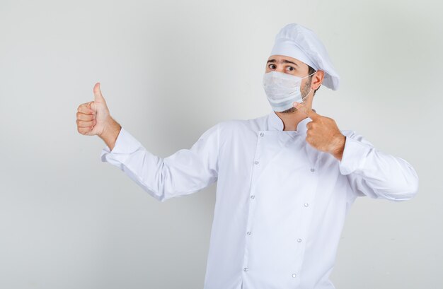 Male chef showing medical mask with thumb up in white uniform and looking careful