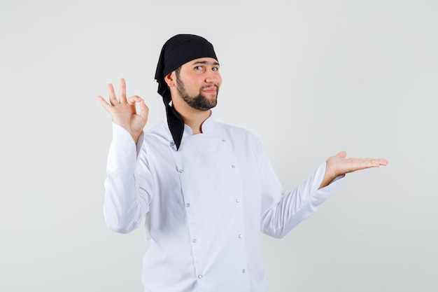 Free photo male chef showing ok sign with palm spread aside in white uniform and looking pleased. front view.