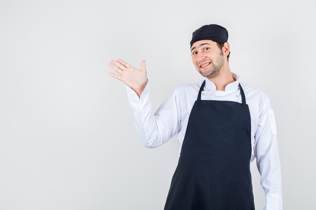 Male chef showing palm to direct visitors in uniform, apron and looking cheerful. front view.