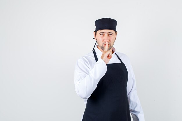 Male chef showing silence gesture in uniform, apron and looking careful , front view.